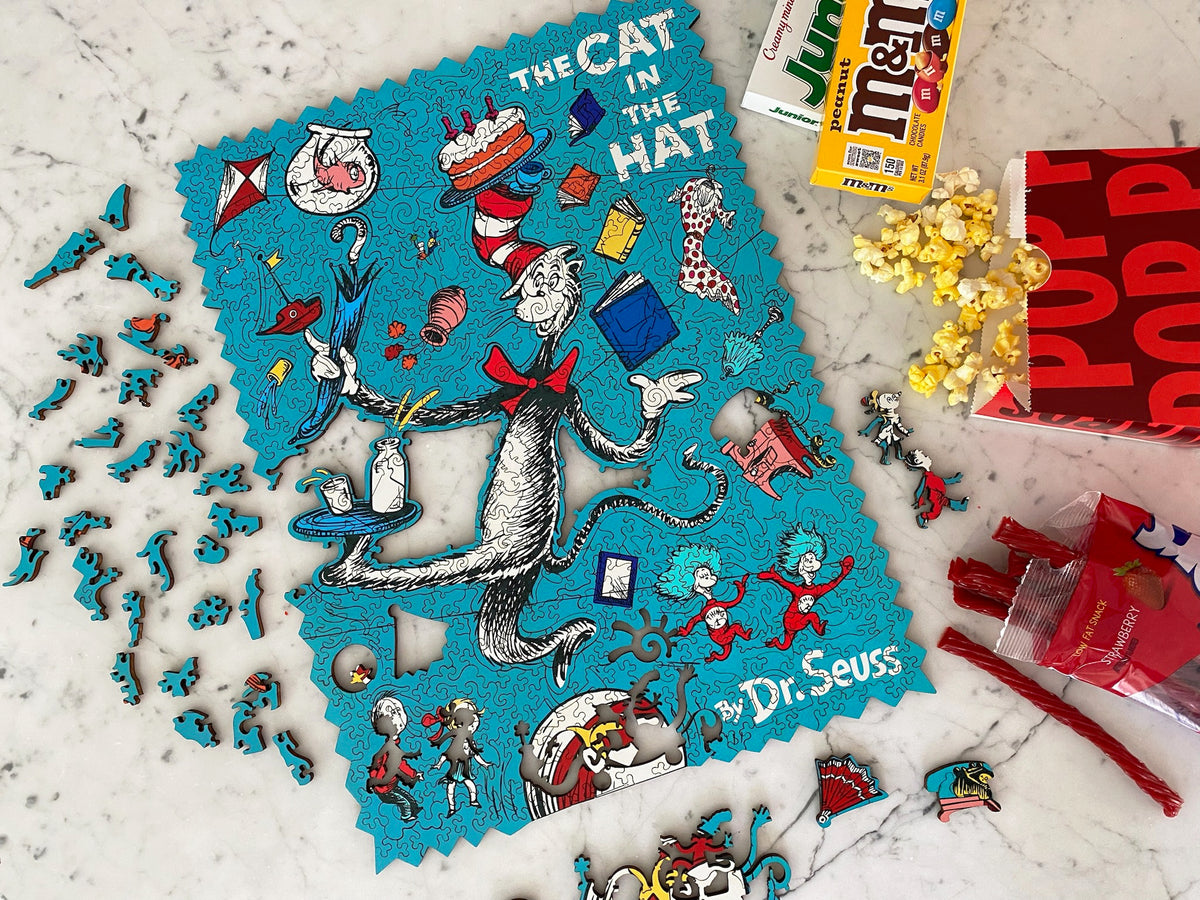 A flat-lay of a Dr. Seuss 'The Cat in the Hat' jigsaw puzzle on a white marble surface, surrounded by themed snacks including M&M's, Reese's, and popcorn.